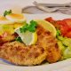 The image shows a Schnitzel decorated on a plate, surrounded by potatoe salad and decorated with a slice of lemon.